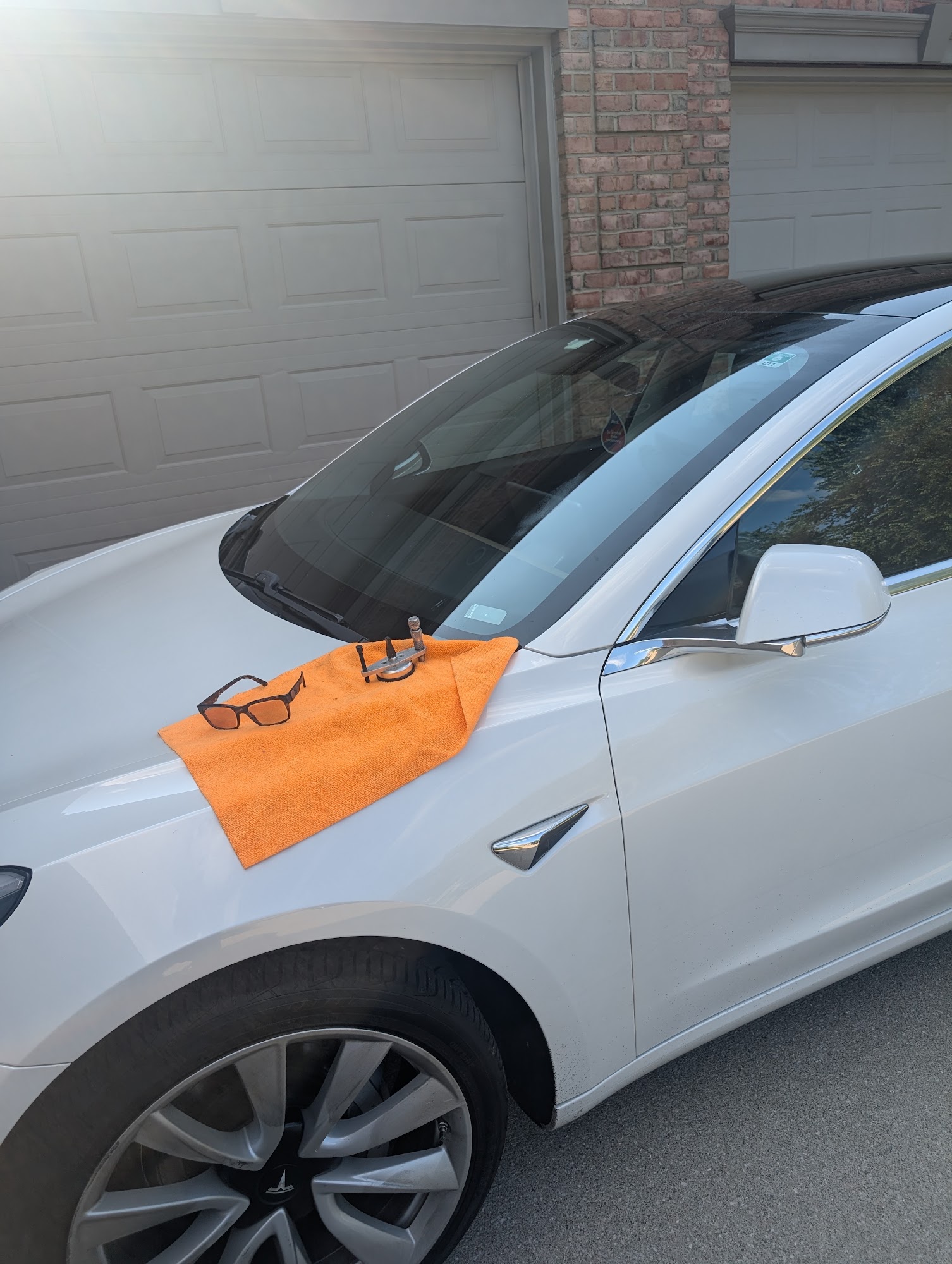 A windshield repair tool clamped to the windshield of a white Tesla parked in a residential driveway, with a microfiber towel on the hood